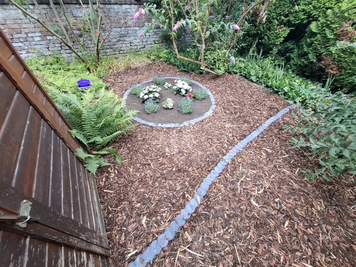 Rundes Pflanzbeet mit Steineinfassung im Rindenmulch, daneben Farne und Stauden